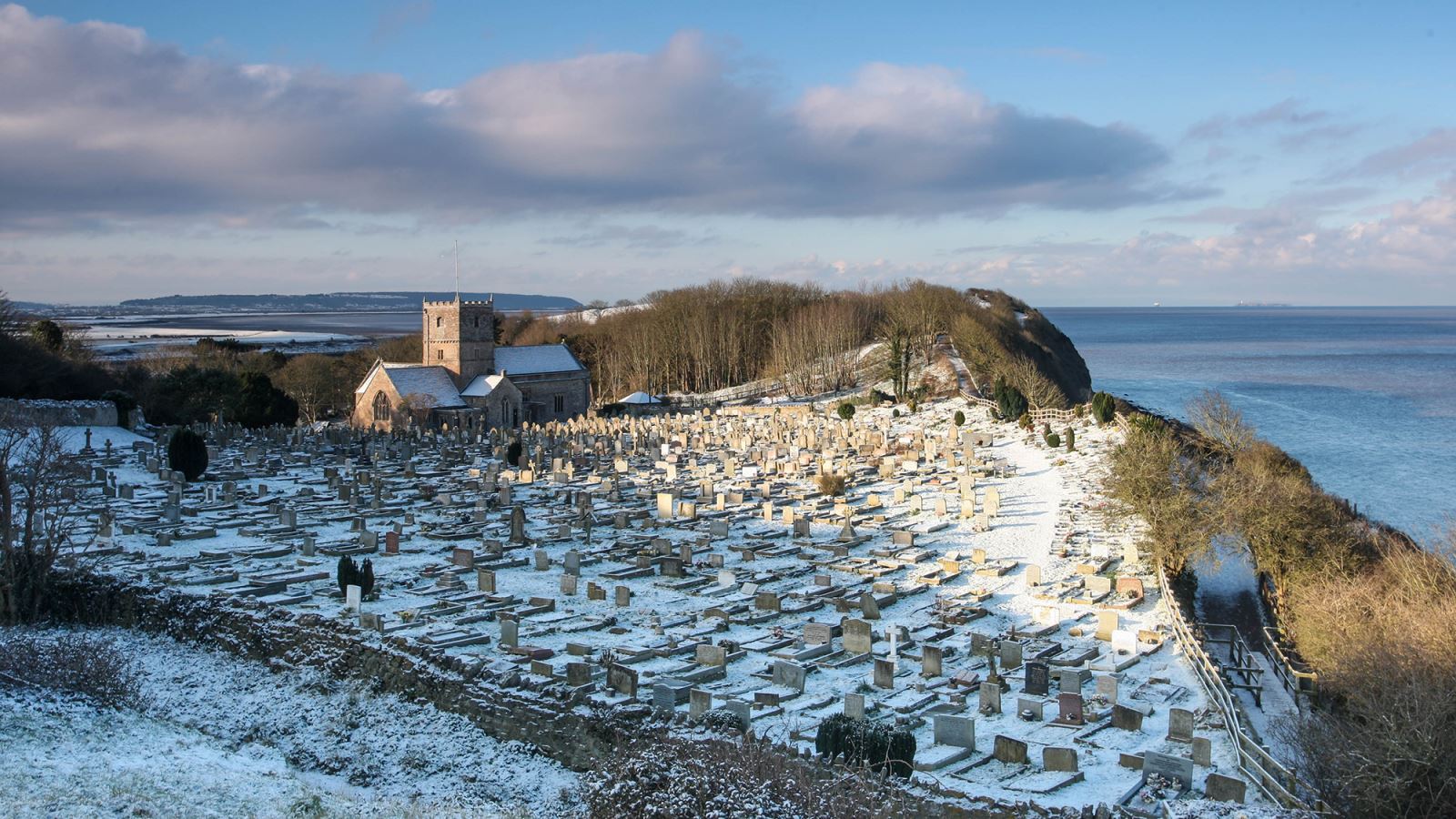 Snowy scene of a church and churchyard overlooking the sea under blue skies